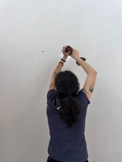 A person wearing a dark-colored t-shirt is using two red and black power drills on a white wall. A thin chain hangs from the wall near the lower drill. The location appears to be indoors, potentially in the Town of Red Hook, United States of America.