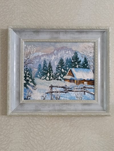 The image shows a framed embroidery artwork hanging on a patterned, light-colored wall. The embroidery depicts a winter scene with a wooden cabin covered in snow, surrounded by snow-covered pine trees, bushes, and a rudimentary fence. A mountain range is visible in the background. The frame is ornate and painted in shades of white and gray. The name "MNA" is embroidered in the lower left corner of the artwork.