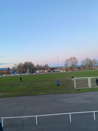The image depicts a soccer field in Karakulino, Russia. The field is adjacent to a paved area marked with white lines. Several individuals are present on the field, including one wearing a blue jacket, one near a soccer goal, and another on the periphery. A soccer goal is visible. The background shows buildings, trees, and a construction crane against a light blue sky.