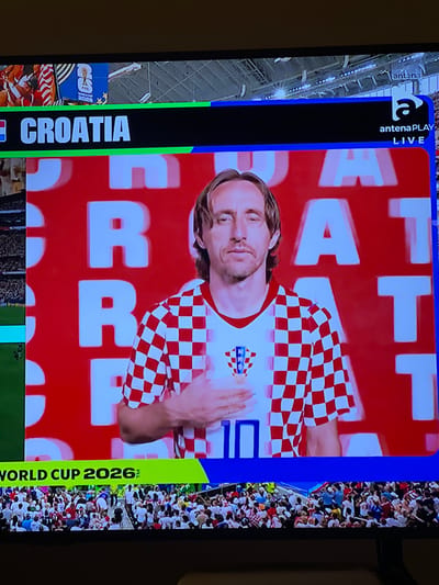 The image shows a television screen displaying a broadcast related to Croatia and the World Cup. A man, likely a footballer, is shown prominently in the center wearing a red and white checkered jersey, the national colors of Croatia. He has his hand on his chest. Behind him, large white letters spell out "CROATIA" and "WORLD CUP 2026". The broadcast also shows logos for "antena PLAY LIVE" and "FIFA WORLD CUP 2020". The bottom of the screen shows a crowd of people watching the broadcast. There is no indication in the image that this scene is located in Florești, Romania.