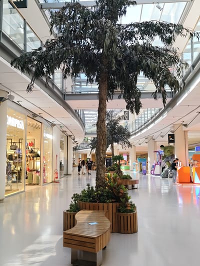 The image depicts an indoor scene within a modern shopping mall, likely the C.C. Auchan Nola in Nola, Italy, given its characteristic architecture and ambiance. In the foreground, a large, artificial palm tree stands in a planter, flanked by wooden benches. The mall features a bright, airy interior with a high, glass-paneled ceiling allowing natural light to filter through. Storefronts line the wide, polished floor, with some shops displaying merchandise and others remaining partially obscured. Several individuals are visible walking or browsing in the background, including a family of three walking away from the camera and other shoppers interacting with displays. A large statue of the Hulk is prominently featured near a gaming or entertainment booth. The overall environment is clean and contemporary, suggesting a busy commercial space.