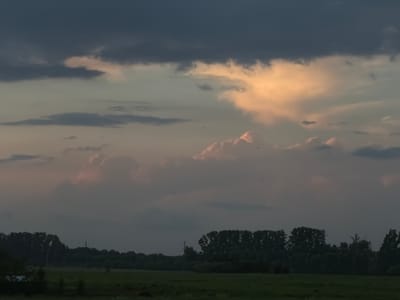 The image depicts a twilight landscape over a grassy field in Leninsk-Kuznetskiy, Russia. The sky is filled with dramatic clouds, with a large dark cloud formation dominating the upper portion. Below this, lighter clouds are illuminated with warm, orange and pink hues, suggesting the setting sun. A band of trees forms a dark silhouette along the horizon, separating the sky from the foreground. The field itself is a muted green, with some darker patches indicating shadows or possibly uneven terrain. There are no visible people or specific objects of interest in the scene. The overall atmosphere is one of natural tranquility at dusk.
