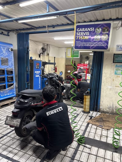 The image depicts the interior of a motorcycle repair shop in Binong, Indonesia. The scene is cluttered with various repair equipment and motorcycles. In the foreground, a person wearing a black shirt with "PIRELLI" visible on the back is crouching down, seemingly working on a black motorcycle. Another person, also in dark clothing, is visible in the background, partially obscured by a blue accordion door, and appears to be working on another motorcycle.

Dominating the background is a blue machine labeled "NITROGEN N4500," a nitrogen tire inflation machine, indicating specialized service. To the left, a large blue vending machine or display unit is present, with "VINFAST" branding. A prominent sign hanging from the ceiling advertises "GARANSI SERVIS 7 HARI" (7-Day Service Warranty), detailing conditions and a website address.

Various tools and equipment are scattered around the floor, including coiled green hoses, a yellow oil drain container, and a piece of cardboard on the floor. The environment appears to be a functional, working repair bay with fluorescent lighting overhead and a tiled floor. The overall impression is one of a busy service center.