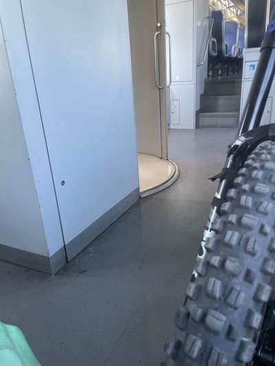 The image depicts the interior of a train carriage, specifically the area near a doorway. In the foreground on the right, the knobby tire of a bicycle is prominently featured, suggesting the bicycle is being transported. To the left, a white wall with a metallic trim at the bottom forms the majority of the scene. A doorway is visible in the middle ground, leading to a slightly raised section of the train carriage. Beyond the doorway, a staircase ascends to a seating area with blue-backed seats. The flooring appears to be a speckled gray material. The lighting suggests an indoor environment, possibly during daylight hours. No people are clearly visible, though the presence of stairs and seating indicates it's a passenger carriage. The image does not provide any specific details about the location being Merichleri, Bulgaria.