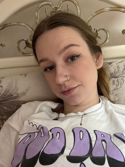 The image depicts a young Caucasian female posing for a self-portrait indoors. She has light brown hair, blue eyes, and fair skin. Her gaze is directed towards the camera with a neutral expression. Visible facial features include a small silver-colored hoop nose ring in her left nostril and a silver-colored ear cuff or multiple small earrings on her right ear. She is wearing a white t-shirt with large, stylized purple lettering, partially legible as "OOD DA", across the chest. A silver-colored chain necklace with a small pendant and an extender chain is visible around her neck. She appears to be reclining or lying down against bedding. The background reveals a light-colored headboard with ornate, possibly gilded, scrollwork detailing. A pillow with a patterned fabric, featuring dark botanical or foliage motifs, is partially visible behind her head and shoulder. There are no identifiable markers in the image to confirm the specific location in the city of Tlustenkhabl, Russia.