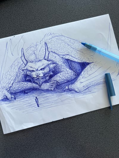 A blue ballpoint pen drawing on white paper depicts a large, dragon-like creature with scales, horns, and a reptilian face, lying down. In front of the creature's head, a small, silhouetted human figure stands. The drawing is set against a dark, textured surface, possibly a table. A light blue pen and its cap are placed on top of the paper. There is no visible indication of the city of Rezina, Moldova.