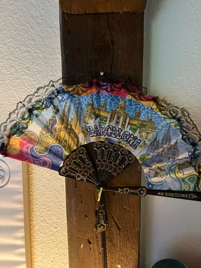 A decorative fan featuring colorful depictions of Barcelona landmarks, including the Sagrada Familia and the Palau Nacional, is attached to a dark wooden beam. The fan is partially opened and adorned with black lace trim. The word "BARCELONA" is visible on the fan. The background consists of a textured white wall and a lighter wooden vertical element. The scene is indoors, with artificial lighting creating shadows. This photograph was taken in Schwäbisch Hall, Germany.