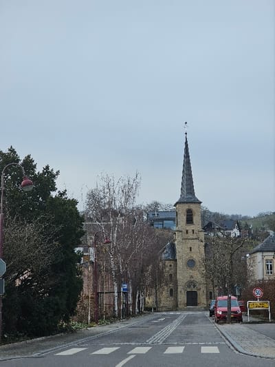 The image displays a street scene in Bech-Kleinmacher, Luxembourg, under an overcast sky, suggesting a cold season due to bare trees. A paved road, featuring a white crosswalk and two central parallel cobblestone strips, extends into the midground, leading to a prominent stone church. The church, constructed from light brown masonry, possesses a tall square bell tower capped with a dark gray, steeply pitched spire adorned with a finial. A circular window is visible on the tower, and the main entrance consists of dark, arched double doors.
On the left side of the road, a dark green, leafy tree and a maroon street lamp are in the foreground. Further along, a row of slender, bare deciduous trees lines the sidewalk, amidst which a blue bus stop sign is positioned. On the right side of the road, a red utility van is parked on the shoulder, adjacent to a yellow sign displaying "Bech-Kleinmacher" and an attached 50 km/h speed limit sign. Residential buildings are scattered on the tree-covered hillsides surrounding the church. No individuals are present within the frame.