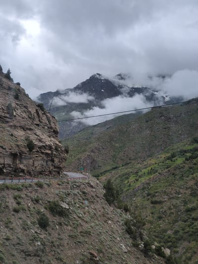 The image shows a steep, winding mountain road in the remote, rugged terrain of Stingri, India. The road, paved and bordered by guardrails, curves sharply to the left as it ascends a rocky, sparsely vegetated slope. In the foreground, a sheer rock face dominates the left side of the frame, its textured surface showing layers of sediment and scattered small trees. The road continues onward and upward, disappearing around a bend.
Further into the distance, the landscape transforms into a series of green, terraced hillsides, indicative of agricultural activity. These slopes are dotted with small trees and shrubs, gradually becoming more rugged and rocky as they meet the foothills of imposing mountains. The peaks of these distant mountains are partially obscured by dramatic, swirling clouds and mist, with patches of snow visible on their upper reaches, suggesting a high-altitude environment.
The sky is overcast with heavy, grey clouds, creating a subdued and dramatic atmosphere. A single, dark power line stretches horizontally across the mid-ground of the image, connecting unseen points and adding a man-made element to the natural landscape. No people are visible in the scene, emphasizing the isolation and vastness of the mountainous region. The overall impression is one of a challenging and awe-inspiring natural environment.