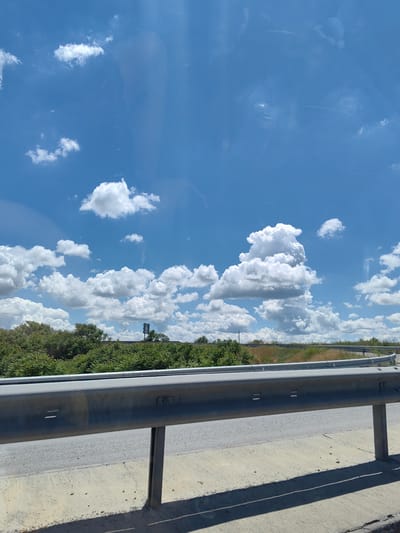 The image captures a view of a highway guardrail in Buhovtsi, Bulgaria, under a bright blue sky dotted with fluffy white clouds. The guardrail, made of weathered metal, runs horizontally across the frame, casting a sharp shadow on the concrete surface below. Beyond the guardrail, a dense line of green trees and shrubs forms a natural barrier, with dry grass visible in the distance. A traffic signal stands prominently among the trees, indicating the presence of a road network. The overall scene suggests a rural or semi-rural highway environment, with the sky and vegetation being the dominant natural elements.