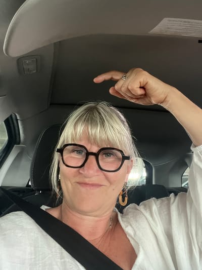 A woman with short blonde hair, wearing black glasses, a white blouse, and a seatbelt, is taking a selfie inside a car. Her left hand is raised and pointing downwards with the index finger extended. The interior of the car is visible, including the headliner, sun visor, and a dome light. The location is not identifiable as Palavas-les-Flots, France.