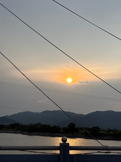 The image captures a serene natural landscape at either sunrise or sunset, viewed from a slightly elevated vantage point. The sky, occupying the upper portion of the frame, transitions from a pale, warm golden hue around the central, luminous sun to a soft, cool greyish-blue towards the edges. The sun, a bright, distinct orb, casts a soft glow, suggesting a hazy atmosphere or thin clouds.
Several dark, thin power lines diagonally traverse the sky, stretching from the upper-left to the lower-right, adding a man-made linear element to the vast expanse.
In the lower half of the image, a calm body of water—likely a river or a lake—reflects the warm light of the sun, creating a shimmering, golden path across its surface. Beyond the water, a dense band of dark green trees or foliage lines the distant bank. A range of majestic mountains, silhouetted in dark blue-grey tones, rises behind the trees, their peaks fading into the atmospheric haze of the horizon.
On the far right, a dark, metallic structure, possibly a part of a railing or a lamppost, stands prominently, partially obscuring the view of the water. A distinctive spherical finial adorns this structure, catching the reflected light from the water. The scene evokes a peaceful and expansive natural setting, characteristic of a heritage area like Nam Natural Heritage Area Trang in Vietnam. No text is visible in the image.