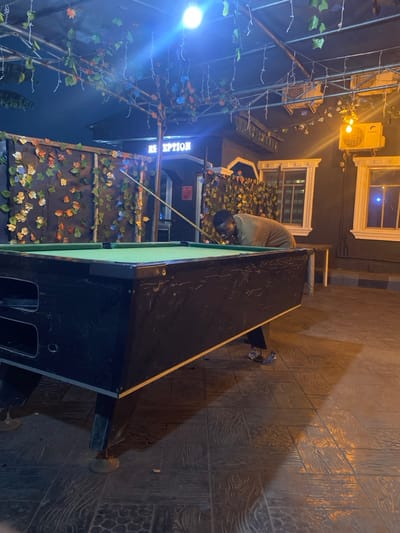The image depicts a nighttime scene at a recreational area in Igbessa, Nigeria. An adult male, dark-skinned and wearing a light brown long-sleeved garment and dark trousers, is positioned at a dark-colored pool table with a light green felt surface. He is leaning over the table, holding a light-colored wooden cue stick with his right hand, preparing to strike a ball. The immediate environment is illuminated by artificial lights, including a bright overhead cool-toned light and warmer-toned string lights in the background. Overhead, a metal trellis structure is draped with artificial foliage featuring green, orange, and brown leaves. A dark-walled building in the background displays a white neon sign reading "RECEPTION" and features two white-framed windows and two white wall-mounted air conditioning units on the right. A partition adorned with similar artificial foliage is visible to the left of the "RECEPTION" sign. The floor is tiled with a textured pattern resembling wood grain. Small debris is visible on the floor near the base of the pool table.