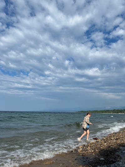 A young female with blonde hair braided down her back walks along a rocky shore. She is wearing a plaid shirt, black shorts with a white pattern, and white sandals. She is looking down at the ground as she walks. The water of a large lake is to her left, with small waves breaking on the shore. The sky above is filled with dramatic clouds. In the distance, a tree-covered shoreline is visible, with mountains beyond. The location is Tolbazikha, Russia.
