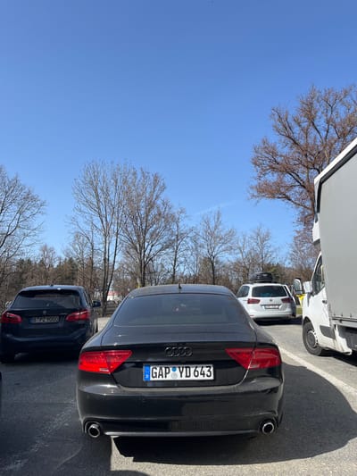 This image depicts an outdoor scene in Icking, Germany, on a clear, sunny day, likely late winter or early spring, as evidenced by the bare deciduous trees in the background. The foreground is dominated by the rear view of a black Audi A7, equipped with dual exhaust pipes and a German license plate displaying "GAP YD 643". To the immediate left of the Audi, a dark-colored BMW, bearing the German license plate "B PZ 9000", is parked. Further back, on the center-right, a white station wagon, possibly a Volkswagen Passat, is visible with a roof-mounted cargo box and its rear driver-side door open. This vehicle displays a Czech Republic license plate, "CZ 484 08". On the far right, the front portion of a white cargo van or small truck is partially visible. The ground is a paved surface, suggesting a parking area or rest stop. No individuals are discernible within the frame.