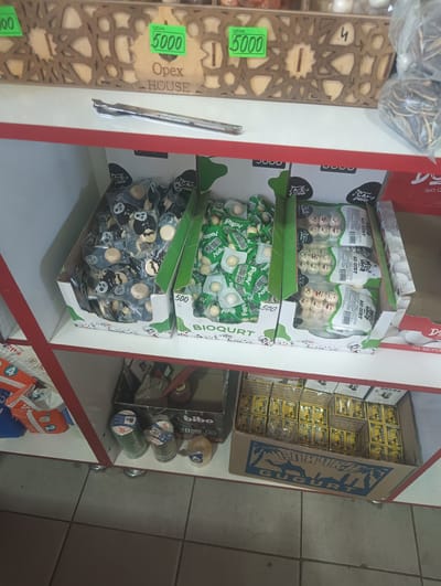 The image shows a retail display shelf in a store in Bulungur, Uzbekistan. On the top shelf, a wooden decorative box with geometric patterns is visible, with two green price tags displaying "5000" in Cyrillic. Below this, on a white shelf, are several cardboard boxes containing individually wrapped candies. One box is filled with candies wrapped in silver foil featuring panda designs, while another box contains candies with green wrappers, some of which show smiley faces. A third box contains candies with white and orange wrappers. On the shelf below, there are spools of tape, including green and blue spools, and a yellow object. A cardboard box with a blue design depicting a camel and palm trees, and the text "СПИЧКИ GUGURT", is filled with smaller boxes, likely containing matches. The overall environment appears to be a small retail establishment with goods displayed on shelves.