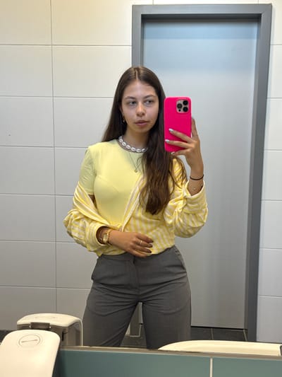 A young woman is taking a selfie in a bathroom. She's holding a pink smartphone up to the mirror with her right hand, with her left hand resting on her midsection. She is wearing a yellow t-shirt underneath a yellow and white striped button-up shirt and gray trousers. She has long brown hair, and is wearing a watch, bracelets, a ring, and a seashell necklace. The bathroom has white tile walls and a gray-framed mirror. A soap dispenser is visible on the right side of the frame. This bathroom is located in Hanau, Germany.