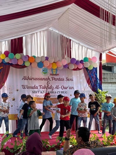 The image depicts an outdoor event, likely a school performance or graduation ceremony, held under a white and maroon tent with hanging decorative ropes. A banner in the background displays text in Indonesian, including "Akhirussanah, Pentas Seni & Wisuda Tahfidz Al-Qur'an" and "SDIT AZ-ZAHRA CURUG". A group of young boys, dressed in casual attire and some wearing caps, are standing on a stage decorated with red carpet and floral arrangements. Some of the boys are actively performing, with one in the center gesturing with his hands and another to his right appearing to sing or speak. The audience is partially visible in the foreground, with one person in a patterned shirt turned away from the camera. Balloons are strung along the top of the stage area. The overall impression is of a vibrant and celebratory occasion taking place in a public or school setting.