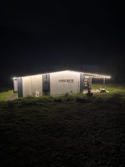A nighttime scene shows a building with "STONE RIVER ECO VILLAGE" written vertically on its side. String lights illuminate the building's edge and a nearby patio area where a person stands. The surrounding environment consists of dark foliage and gravel. The location is presumed to be Kapreshumi, Georgia.
