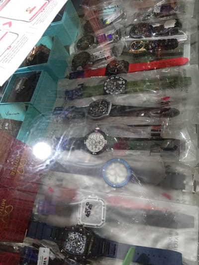 This image shows a close-up, eye-level view of a display of watches, likely in a store or market stall in Gampongteungoh, Indonesia. The scene is brightly lit, possibly from overhead lighting reflecting off the plastic packaging of the watches. Various watches are neatly arranged, some with visible straps in red, green, and blue. Many of the watches are in clear plastic packaging, displaying their faces and bands. Some packaging includes text such as "WATER PROOF FASHION WATCH" and "ORIGINAL Water Resist". There is also a red box with the word "MIRAGE" and a logo. On the left side of the image, a portion of a document or flyer is visible with some text in what appears to be Indonesian. The overall impression is of a commercial setting showcasing a variety of affordable or fashionable timepieces. The focus is entirely on the merchandise, with no people clearly visible. The arrangement suggests a retail environment where these watches are for sale.