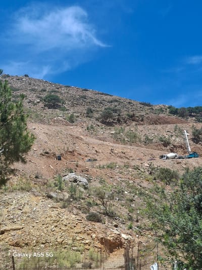 The image shows a steep, rocky hillside under a bright blue sky with wispy clouds. A concrete mixer truck is parked on a cleared path partway up the slope. There is equipment with an articulated boom extending from the truck. The surrounding terrain is covered in sparse vegetation, and a fence is visible on the right edge. It is not possible to independently determine if this location is in Gazi Municipal Unit, Greece, without additional information.