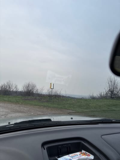 The scene depicts an outdoor landscape in Gornik, Bulgaria, viewed from inside a vehicle. The upper two-thirds of the image show an overcast, light grey sky. Below, a green grassy terrain with scattered leafless trees and bushes extends, featuring patches of brownish-purple vegetation. A dirt or gravel path is visible in the lower-left foreground. A yellow and black horizontally striped rectangular signpost stands amidst the bare bushes in the mid-right. A hazy distant horizon is faintly visible through the vegetation. The vehicle's dark grey dashboard occupies the lower portion of the frame, with windshield wipers at the base of the windshield. A partial view of a black rearview mirror is in the upper-right. The windshield displays reflections, including a rectangular light reflection in the upper-middle. A white packet, possibly a snack or wipes, with colorful graphics and the partial word "Jussy," rests in a dashboard compartment. No individuals or specific actions are discernible.