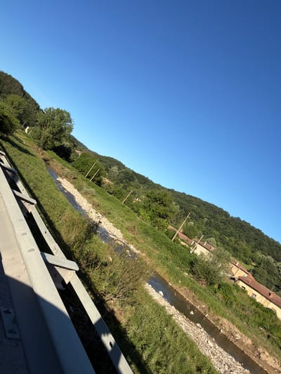 The image is a landscape photograph taken from a slightly elevated position, possibly a bridge or overpass, looking down onto a rural scene in the city of Veselinovo, Bulgaria. The photograph is tilted, with the sky occupying the upper right portion of the frame and the ground filling the lower left.
In the foreground, a metal guardrail runs diagonally from the bottom left corner upwards and to the right. It is constructed of thick, grey metal bars with visible bolts and rivets. The guardrail casts a distinct shadow across the grassy embankment to its right.
Beyond the guardrail, a shallow, rocky stream flows from the background towards the foreground. The stream bed is composed of various sizes of grey and brown stones. The banks of the stream are covered in dense green grass and interspersed with small shrubs and weeds.
The terrain slopes upwards from the stream, forming a grassy hill. Scattered along the hillside are several poles with electrical insulators, suggesting utility infrastructure. Further up the slope, there are clusters of trees and patches of green vegetation.
In the middle ground and background, several buildings with terracotta-tiled roofs are visible, nestled amongst the trees and on the hillside. These appear to be traditional rural homes. The architecture suggests an older, possibly rustic style.
The entire scene is bathed in bright, natural sunlight, indicating it was taken during the daytime. The sky is a clear, vibrant blue, with no clouds visible. The overall impression is one of a serene, sunny day in a hilly, rural area.
