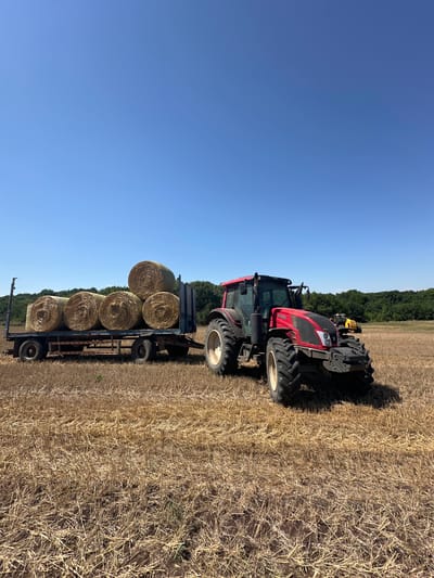 In Bistrets, Bulgaria, on a clear, sunny day, a red Valtra tractor is parked in a harvested field, with the stubble of cut crops covering the dry, brown earth. The tractor is positioned to the right of the frame, facing slightly towards the left, its large, rugged tires casting shadows on the ground. Attached to the rear of the tractor, and extending to the left, is a trailer laden with five large, round hay bales. The bales are stacked in two layers, with three in the bottom layer and two on top, their tightly wound straw a golden-yellow hue. Behind the trailer, a line of dense green trees forms a natural border against the bright blue sky, suggesting the edge of a forest. Further in the background, to the right of the tractor, another yellow piece of farm equipment, possibly another tractor, is visible in the field. The overall scene depicts agricultural activity, likely the collection of hay after harvest.