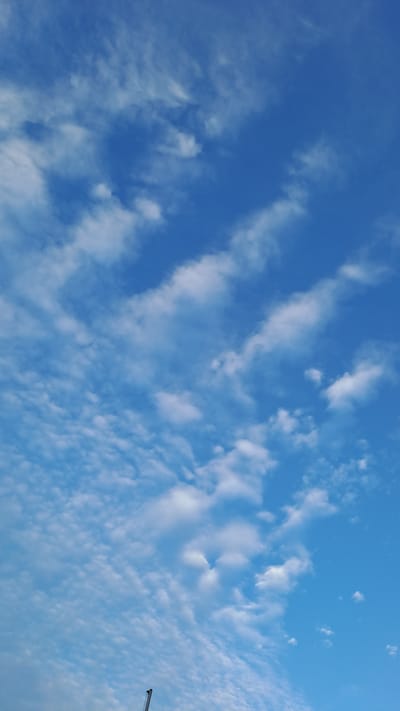 A bright blue sky fills the frame, adorned with a variety of white clouds. In the lower center of the image, a portion of a dark, slender object, possibly part of a crane or antenna, is visible against the sky. The clouds are dispersed, with some appearing as wispy streaks, while others are more rounded and fluffy, creating a dynamic texture across the expanse of the sky. The overall impression is one of a clear, breezy day.