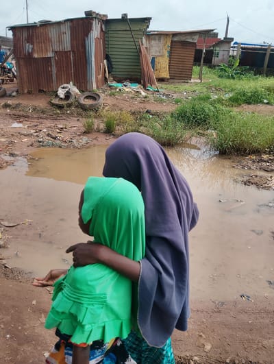 This image depicts a scene in Orudu-Olokan, Nigeria, focusing on two individuals, presumably a mother and child, standing in a muddy, debris-strewn area. The child, wearing a vibrant green headscarf and dress with ruffled layers, is embraced by an adult figure draped in a dark purple headscarf and cloak. The adult's arm is around the child's shoulder, and the child's left hand is extended slightly.
The environment is characterized by a large, murky puddle of water dominating the foreground, reflecting the overcast sky. The ground is predominantly dirt, mixed with scattered trash, including plastic debris and what appear to be discarded fabric. In the background, ramshackle structures built from corrugated metal and wood suggest a makeshift settlement or informal housing. Some of these structures show signs of rust and wear. Two old tires are visible near one of the buildings. Lush green grass grows in patches throughout the area, interspersed with the mud and refuse. The overall impression is one of poverty and difficult living conditions.