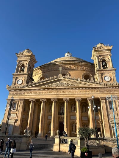 The image depicts the Mosta Rotunda, officially the Basilica of the Assumption of Our Lady, located in Mosta, Malta. The scene is presented under a clear, bright blue sky, with the church facade illuminated by direct sunlight originating from the left, casting shadows to the right.
The grand Neoclassical church dominates the frame, featuring a large circular dome topped by a smaller cupola. Its front facade comprises a prominent portico with six Corinthian columns supporting a triangular pediment. Above the columns, an entablature bears a Latin inscription: "VIRGINI SYDERIBVS RESTITVTAE. T. H. MVSTENSES FF. A. S. M. CCM. LVII." Symmetrically flanking the main structure are two bell towers, each with an arched opening containing a bell and a circular clock face. The clock on the left tower indicates approximately 10:20, and the clock on the right tower shows approximately 10:15. Decorative niches containing statues are visible between the columns and on the upper levels of the bell towers. Intricate carvings adorn the architecture.
In the foreground, a paved plaza precedes a wide set of stone steps leading to the church entrance. Approximately ten individuals are present: A group of three (two females, one male) stands on the lower left, facing partially away. A person in a grey hoodie ascends the steps. Another individual in a dark jacket and backpack walks away from the church on the right. Several other figures are scattered on the steps and near the entrance. Two ornate black streetlights with multiple globes are positioned on the plaza. On the right, a tall, vertical metal structure adorned with white lights, possibly seasonal decorations, stands near a low stone railing. A small olive tree in a dark planter, featuring a shield emblem with a red cross on a white background, is situated on the right foreground.
