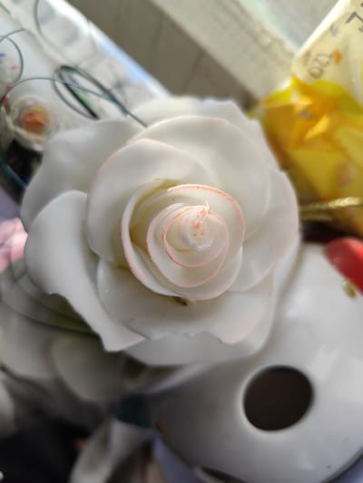 This close-up, vertical, high-angle shot focuses on a white ceramic rose, possibly a decorative item. The rose has delicate pink accents along the edges of its inner petals, creating a subtle gradient. The petals are layered and appear smooth and glossy.
In the background, out of focus, is a bright yellow object, possibly wrapping or a toy, with some faint text visible. The setting appears to be indoors, near a window, suggested by the soft light illuminating the scene. The lighting indicates it could be daytime, with natural light casting soft shadows and highlights on the ceramic rose. There are also some thin, green wires visible in the upper left portion of the image, perhaps part of the rose's decorative stem or another surrounding object. The overall mood is calm and serene, with the focus on the intricate details of the rose. The location context of Yenakiieve, Ukraine, does not provide further visual cues within the image itself.