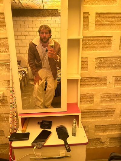 The image displays an indoor scene within a room, identified as being in a salt hotel in Uyuni, Bolivia, evidenced by the distinctive construction of the walls from textured, rectangular blocks of a light-colored granular material, consistent with salt.
A male individual is observed in a full-length mirror, actively taking a selfie with a light-pink cased smartphone held in his right hand. He stands with a slight forward lean and bent knees, his left hand resting on his left thigh. The subject has short, light-brown hair and a beard. He is dressed in a brown jacket with a white shearling collar and inner lining, a white crew-neck t-shirt, and light-beige cargo pants. A light-grey cross-body strap is visible across his torso, and a light-colored beaded bracelet adorns his right wrist.
The mirror, framed in white, is integrated into a white and pink dresser-vanity unit. This unit includes two open shelves positioned to the right of the mirror. On the dresser's surface, below the mirror, a dark-colored smartphone (consistent with an iPhone 14 Pro/Pro Max) is charging via a white cable connected to a black portable power bank. Also present are a black JBL Clip 4 portable Bluetooth speaker, a black paddle hairbrush, a clear, rectangular glass perfume bottle, and an orange-wrapped cylindrical object.
The background visible in the mirror's reflection reveals a bed with striped bedding (dark and light blue), a light-colored wall socket with a white plug, and a vertical fabric strip adorned with multicolored circular patterns. To the right of the dresser unit, the salt-block wall construction continues, forming the dominant environmental feature. The scene is illuminated by warm-toned lighting, imparting a yellowish cast.