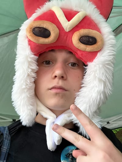 A young person, with light skin and brown eyes, wears a red, plush hat resembling an animal or character with white fuzzy trim. The hat has two large, oval-shaped eye designs with dark brown centers and light brown rims. The person is making a peace sign with their right hand, index and middle fingers extended. They are wearing a black t-shirt and a blue and white checkered scarf or strap is visible over their left shoulder. A round, blue and silver pin is attached to the t-shirt. The background appears to be a green tent or tarp. The image seems to be taken outdoors, possibly in a park or at an event. This image was captured in Nagakute, Japan.