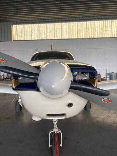 A single-engine propeller aircraft is positioned centrally within a hangar at Toussus-le-Noble Airport, Toussus-le-Noble, France. The aircraft, facing the viewer, is predominantly white with a dark blue upper fuselage section detailed with gold pinstriping. Its two-bladed propeller, equipped with a silver spinner, features a "HARTZELL" logo on the visible blade. The front landing gear, with a red rope or tow bar attachment, is in the foreground. Engine components, including cylinders, are visible through an intake opening on the right side of the cowling. The hangar environment consists of a dark concrete floor, white walls, and a translucent corrugated roof section admitting ambient light. In the background to the right, a red step ladder and several white cylindrical containers are present. No individuals or active interactions are visible.