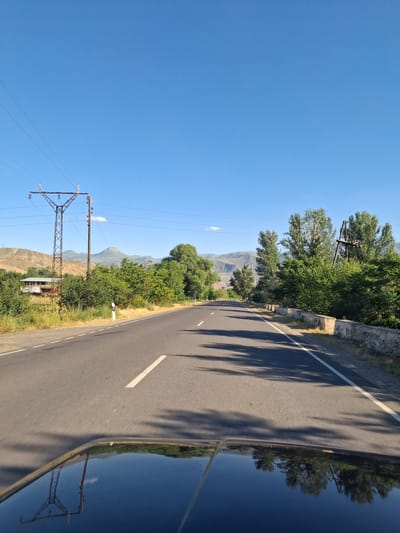 The image depicts a road in a rural, mountainous area, likely in Getap, Armenia. The sun is shining brightly, casting long shadows from trees onto the asphalt road. The sky is a clear, deep blue, suggesting a pleasant, sunny day. Lush green trees and vegetation line the side of the road, contrasting with the dry, brown hills in the background. A power line tower stands prominently on the left side of the road, with wires stretching across the frame. The road itself is paved and has a white dashed line marking its center. The reflection of the sky, trees, and power lines is visible on the dark, glossy surface of what appears to be a vehicle. The overall scene conveys a sense of peaceful travel through a scenic landscape. There is no visible text in the image.