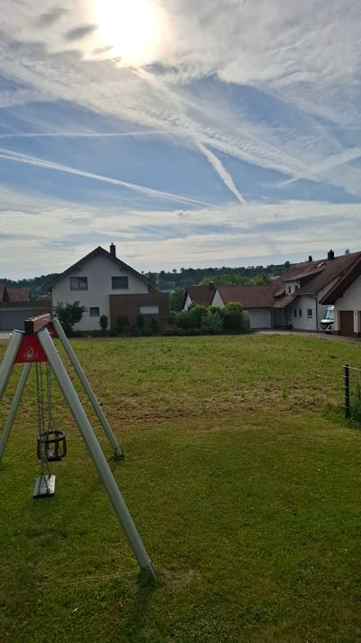 A wide, eye-level, outdoor, daytime, eye-level shot shows a suburban residential area in Buchen (Odenwald), Germany. In the foreground, a metal swing set with a single plastic seat is positioned on a grassy lawn. The swing seat is dark red. Beyond the lawn, several residential houses with red tile roofs and white or light gray walls are visible. Some of the houses have attached garages. A white van is parked in front of one of the houses. A black metal fence is partially visible on the right side of the frame. The sky is blue with scattered white clouds and numerous white contrails, suggesting airplane activity. The sun is visible in the upper left portion of the sky, partially obscured by clouds. The overall scene depicts a quiet, residential neighborhood on a clear day. There are no people or animals visible in the image.