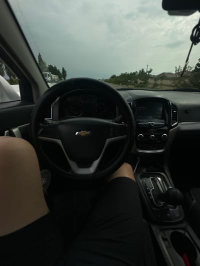 The image is taken from the perspective of someone driving a car. The dashboard, steering wheel with a Chevrolet logo, gear shift, and a person's legs are visible. Outside the car, there are trees, houses, and a cloudy sky. In the distance, a truck is visible. The scene appears to be in Sergeli, Uzbekistan, based on the provided information. The weather is overcast.