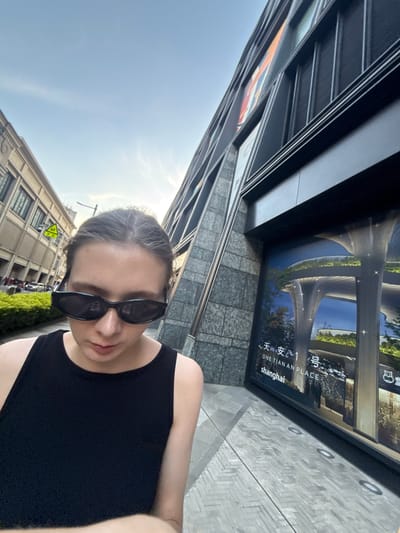 The image captures an eye-level, outdoor selfie-style shot of a person in an urban environment, likely in Putuo District, Shanghai, China.

The central subject is a person, appearing female, shown from the chest up, wearing a simple black sleeveless top and dark, rectangular-framed sunglasses. Her hair is pulled back, and she is looking down towards the camera. Bright sunlight illuminates her forehead and parts of her face.

The setting is a city street or public square on a clear, sunny day. To the person's right, a large display or billboard is mounted on a modern building. This display features an architectural rendering of a contemporary complex with curvilinear structures and green landscaping. Visible text on the display includes "天安 1" (Tian An 1), "ONE TIAN AN PLACE," and "shanghai," indicating the project and its location. The ground in front of the building is paved with light-toned, patterned tiles, interspersed with circular embedded lights.

In the background to the left, another building with a more traditional architectural style, characterized by numerous windows, stands against a clear blue sky. Below this building, there's visible greenery, suggesting bushes or small trees, along with parked vehicles and a yellow triangular warning sign with a black symbol. The scene depicts a vibrant urban area under bright daylight conditions, showcasing a blend of architectural styles.
