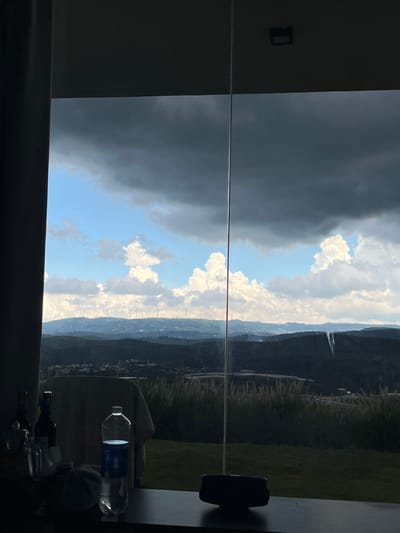 The image shows a view from inside a building, looking out through a large window or glass partition at a landscape. In the foreground, on a dark surface, are several objects: two dark-colored bottles, a clear glass object, a plastic water bottle with a blue label, and a dark-colored item that appears to be a small electronic device. A light-colored cloth is draped over what looks like a chair behind these objects.

The background reveals a rolling landscape with dark green hills and some buildings visible in the distance. On the ridge of one of the hills, several wind turbines are lined up. The sky is partly cloudy, with large, dark storm clouds dominating the upper portion of the frame, contrasting with patches of bright blue sky and illuminated white clouds. A thin, vertical line, likely a seam in the glass or a structural element of the window, divides the view. The scene appears to be set in a location with a somewhat dramatic sky, possibly indicating an approaching storm. The mention of Đa Lộc, Vietnam, suggests a specific geographical context for this landscape.
