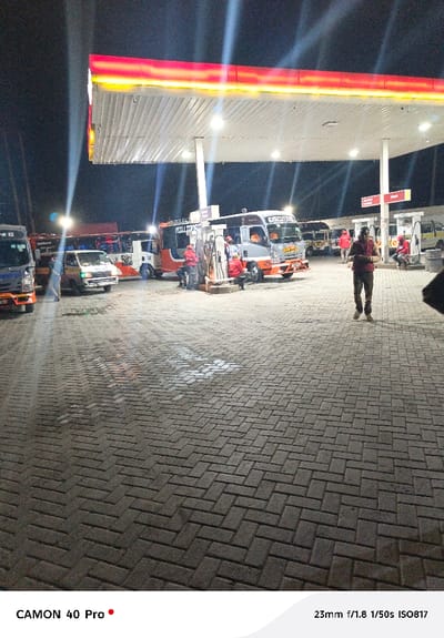 The image captures a nighttime scene at a bustling gas station, illuminated by bright overhead lights under a wide canopy. The canopy itself features prominent red and yellow trim along its edges. The ground is paved with dark interlocking brick pavers laid in a herringbone pattern, showing some wet patches or reflections of light.
Several vehicles, predominantly minibuses or vans, are present at the pumps. On the far left, part of a blue minibus is visible. Closer to the center-left, a light-colored van/minibus with people inside and around it is parked. Further right, a grey and orange minibus, with "EXECUTIVE" visible on its side, is positioned near a fuel pump. Another similar orange and grey minibus with text that appears to read "MILLTON..." on its front is also near a pump. In the background to the right, more vehicles can be seen.
Gas station attendants, identifiable by their red uniforms, are actively engaged in fueling the vehicles. Several other individuals, likely drivers or passengers, are also standing or sitting around the pumps and vehicles. A prominent figure, a man in a dark red shirt and dark trousers, stands towards the right side of the frame, appearing to look at his phone. Another person's shoulder and arm are partially visible in the bottom right corner, indicating the perspective of the photographer.
The bright artificial lighting creates distinct starburst effects around the light sources, contrasting with the dark night sky. Signs for "Diesel" are visible above some of the fuel pumps. The overall atmosphere suggests a busy late-night or early-morning stop for vehicle refueling.
Metadata visible at the bottom of the image indicates it was captured with a "CAMON 40 Pro" camera, with settings of "23mm f/1.8 1/50s ISO817".