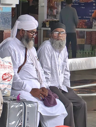 Two men, appearing to be in their late 50s or older, are seated outdoors. The man on the left wears a white turban and a white tunic, with a brown strap draped over his shoulder. He has a gray beard and glasses. The man on the right wears a black cap and a white patterned shirt, also with a gray beard and glasses. They are seated next to each other on a bench, with luggage beside them. The background suggests a market or a public transport waiting area, with shelves of goods and people in the distance. The image seems to have been taken in Khandwa, India, indicated by text on a nearby bag.