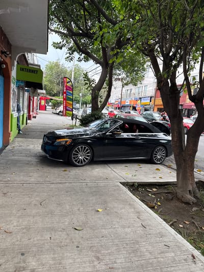 A black convertible Mercedes-Benz C-Class is parked on a sidewalk in front of a building with shops in Tizapan, Mexico. The car's top is down, revealing red interior seats. The sidewalk is made of concrete and is partially covered with fallen leaves. A person in a gray hooded sweatshirt and blue jeans is standing outside one of the shops, looking into the building. To the left of the car, a green sign reads "INTERNET". A colorful vertical banner with text in Spanish, including "CENTRO DE COPIADO", "IMPRESIONES", and "PAPELERIA", is visible further down the sidewalk. Trees with green leaves are growing along the sidewalk and in front of buildings across the street. The street is lined with buildings, some of which appear to be businesses. Cars are parked and driving on the street in the background. The sky is overcast.
