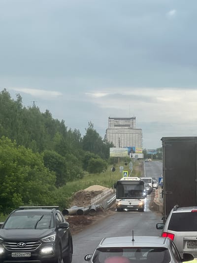 The image captures a traffic jam on a road likely within or near Новосёлки, Russia, under an overcast sky. In the foreground, a dark grey Hyundai SUV is visible, its license plate reading "0822EH 792". To its right, a portion of a white SUV's rear is seen, with a license plate partially obscured but showing "K04". Further ahead, a dark-colored truck is stopped, followed by a white bus with illuminated headlights. Several other vehicles are also present in the queue.

The roadside features construction activity, with large pipes laid out and piles of sand and dirt. A concrete barrier runs along the side of the road. In the background, a large, multi-story building with a light-colored facade dominates the skyline. Adjacent to the road, billboards are placed, one featuring an image of a person in uniform and text in Cyrillic script, another with the word "ТОН". Dense green trees line the left side of the road, extending into the distance. A power line tower can be seen rising above the treeline on the left. The road surface appears wet, suggesting recent rain.