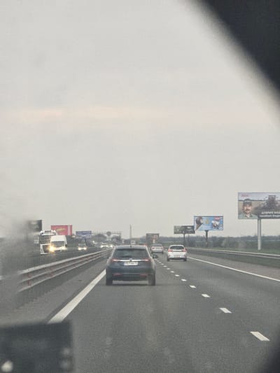 The image depicts a multi-lane highway scene in Opalchenets, Bulgaria, observed from inside a vehicle traveling forward under an overcast sky. The dark asphalt road occupies the majority of the lower frame, featuring white solid and dashed lane markings. A gray barrier system lines the left side of the highway.
In the mid-ground, a dark grey hatchback, bearing Bulgarian license plate "PB 7166 TX", is prominently visible directly ahead in the center lane. Several other vehicles, including cars, vans, and a white semi-truck with its headlights illuminated, are visible further down the road in both lanes, all moving in the same direction.
The environment flanking the highway consists of flat, vegetated terrain with distant trees. Multiple large billboards are erected along the right side of the road. The most distinct billboard on the far right displays a man with a mustache and cap next to a tractor, advertising "NIK" (likely agricultural machinery, with Bulgarian text "ДОБРИТЕ МАШИНИ" - Good machines and "посяват бъдеще" - sow the future). Another billboard features an ice cream or dessert product.
The upper right and lower left portions of the image are partially obscured by elements of the capturing vehicle's interior, and a slight blur or smudge is present on the left side of the windshield.