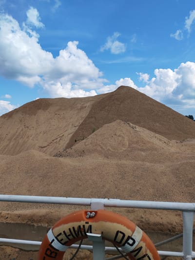 In Vasil'ki, Russia, a large, man-made sand pile dominates the landscape under a bright blue sky with scattered white clouds. The sand pile appears to be a significant construction or industrial material deposit, with various slopes and formations. In the foreground, a white railing runs horizontally across the image, suggesting a viewing platform or the edge of a structure. A life preserver, orange and white with visible Cyrillic lettering, is positioned to the right of the railing, partially obscuring it. The overall scene conveys an industrial or operational setting related to material handling or construction in Vasil'ki.
