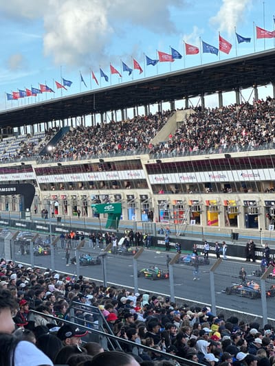 This image depicts a motorsport event at a race circuit in Le Mans, France, viewed from a spectator grandstand. The upper section of the image shows a partly cloudy sky above the grandstand structure. Along the top edge of a dark, elevated grandstand roof, approximately 20-25 flags are displayed, primarily alternating between red and blue, with some bearing a white "GP Explorer" logo.
Below the roof, multiple tiers of concrete grandstands are densely populated with thousands of spectators, all facing towards the track. Beneath the main grandstands, a row of corporate boxes features large windows and "Boursorama Bank" signage.
The mid-ground reveals the pit lane area, comprising several open garage bays with numbered markings (e.g., "1," "2," "3"). Branding such as "Samsung Galaxy A" and "GP Explorer" is visible on the pit lane walls and structures. Numerous individuals, identified as race personnel or officials, are present in the pit lane, some standing near the garage entrances. A tall metal scaffolding is positioned near pit box 2.
The immediate foreground is occupied by the backs of several dozen spectators in a lower grandstand section, positioned behind a metal safety fence. These spectators are observing the asphalt racetrack, which extends horizontally across the lower middle of the image.
On the racetrack itself, multiple open-wheel race cars are visible on what appear to be grid positions, marked by white lines. From left to right, a green and black car, a light blue and black car, and a dark blue and orange/red car (bearing Red Bull livery) are clearly identifiable, positioned on the track, suggesting a pre-race or pre-session scenario.