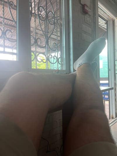 The media file shows a low-angle, close-up shot of a person's legs and feet, wearing socks, propped up against a window frame. The scene appears to be indoors, with a glimpse of an outdoor environment visible through the window.
**Subjects:** The primary subjects are the legs and feet of a person. The person is wearing light gray socks. The background shows parts of a window frame, some white rectangular objects that could be light switches or electrical outlets, and a red object that resembles a fire alarm or emergency button. Outside the window, there are some metallic bars, a corrugated metal roof, and greenery, suggesting an outdoor space.
**Activity or Event:** The person's legs are resting, indicating a moment of relaxation or pause. The focus is on the lower body, suggesting the shot might be from the person's own perspective.
**Notable Details:** The lighting suggests it is daytime, with sunlight streaming through the window, creating some lens flare and washing out certain areas of the image. The metallic bars outside the window are ornate, possibly decorative security bars. The white objects on the wall are modern in design. The presence of a fire alarm or similar device indicates a functional space, possibly a home or a commercial building. The text "CK" is visible on the sock.
**Location Cues:** The user-provided location context is Nikopol, Ukraine. While the image itself doesn't offer definitive geographical clues, the presence of what appears to be security bars on the window, combined with the general setting, could be consistent with environments in Ukraine.
**Emotions:** The image conveys a sense of casualness and perhaps a moment of quiet observation from a relaxed posture.