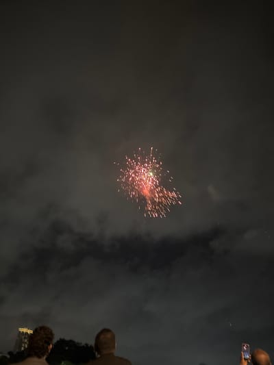The image captures a night scene in Sri Jayewardenepura Kotte, Sri Lanka, featuring a fireworks display against a cloudy sky. In the foreground, the backs of three people are visible, looking up at the sky. The person on the right is holding up a mobile phone, seemingly recording or photographing the event. In the background, a tall, illuminated building is partially visible, indicating an urban setting. The fireworks are a cluster of red and white lights, arcing and dissipating in the dark sky, with smoke trails indicating their recent detonation. The overall atmosphere suggests a celebratory public event.