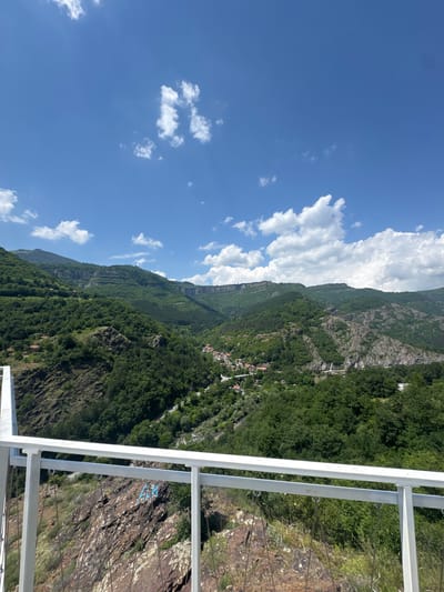 This image displays a panoramic outdoor scene during daylight hours, observed from an elevated vantage point in Zasele, Bulgaria. A white metal railing occupies the foreground, from which thin cables descend towards a steep, reddish-brown rocky incline below. Blue spray-painted Cyrillic letters "АК" are visible on this rock face in the lower-left quadrant. The mid-ground and background are dominated by a vast expanse of green, densely forested mountains and valleys. Prominent rocky cliffs and layered geological formations are visible along the mountain ridges. A small settlement featuring houses with orange roofs and a visible road and bridge system is nestled within a central valley. The sky is clear blue with scattered white cumulus clouds. No individuals are present in the frame.